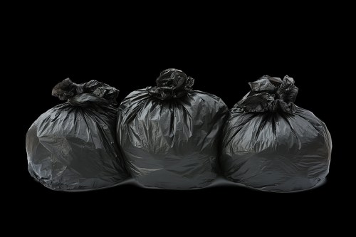 Three large black plastic rubbish bags, tightly knotted at the top, are arranged in a row on a dark surface against a black background. The bags appear full and are made of glossy, slightly crinkled plastic material, with visible creases and folds. They are evenly spaced, with the bag on the left slightly leaning back and the middle bag standing upright, while the right bag sits directly on the surface. The setting suggests an outdoor or warehouse environment, possibly at a commercial waste removal site in Holborn, with no additional objects or environmental context visible. The image emphasizes the uniformity and urgency of rubbish collection, relevant to a professional rubbish removal service such as Commercial Waste Holborn, which specialises in handling waste from businesses or properties in the local area.