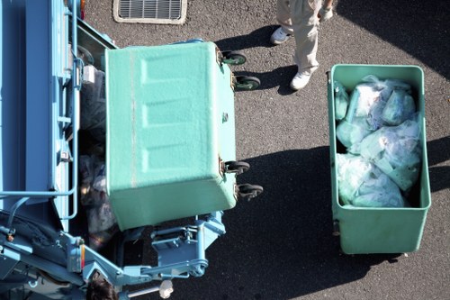 An overhead view of a green wheeled rubbish bin placed on a paved surface, filled with clear plastic bags containing assorted waste materials, with part of a blue refuse collection vehicle visible on the left side. The scene appears to be outdoors, possibly on a driveway or service area, with the lower half of a person wearing beige trousers and visible shoes standing nearby. The environment and waste items suggest a rubbish removal or waste collection operation, typical of commercial waste services in Holborn, London, with the focus on efficient collection and disposal of household or business waste.