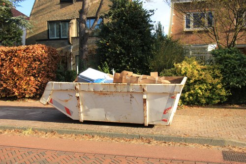 A large, white, industrial-style skip filled with cardboard boxes and miscellaneous waste materials, positioned on a paved residential street pavement near the edge of the road. The skip's exterior shows signs of rust and dirt, with visible red and white reflective safety markings on its sides. Surrounding the skip are residential houses with brick and weatherboard exteriors, set amidst trees and shrubbery, indicating a suburban area. The background features a mix of greenery including a tall, dark green bush and several deciduous trees with autumn foliage, suggesting an early autumn season. The pavement beneath the skip is a combination of brick and concrete, with a curb separating it from the road, which is lined with similar street features. The scene is lit by natural daylight, casting shadows from the trees and the skip itself, and is representative of rubbish removal activities conducted by companies such as Commercial Waste Holborn in the central London boroughs.