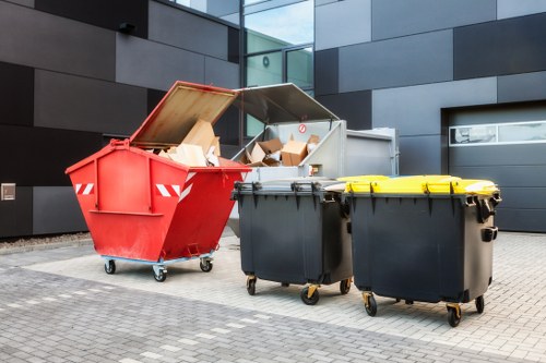 The image shows three large wheeled waste containers situated outdoors on a paved area adjacent to a modern building with dark grey and glass cladding. The first container on the left is a red skip with an open top, filled with various cardboard boxes and packaging waste; its lid is propped open, revealing the contents inside. To the right are two black wheelie bins with yellow lids, positioned side by side, and closed. The scene appears to be part of a commercial waste collection area, fitting for a rubbish removal service in the Holborn area, with the containers used for collecting and disposing of commercial or industrial waste. The environment is clean and well-maintained, with the building’s reflective glass surfaces and modern architectural design in the background, suggesting an urban setting suitable for businesses requiring regular waste management services. The composition emphasizes the different types of waste containers used by Commercial Waste Holborn for efficient rubbish collection and disposal in central London’s commercial districts.