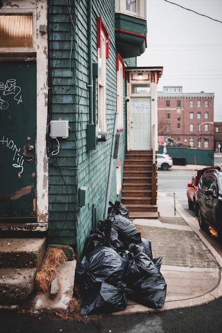 A collection of black plastic rubbish bags filled with waste, stacked against the green exterior wall of a building in an urban setting. The wall features weathered, dark green wooden shingles with so