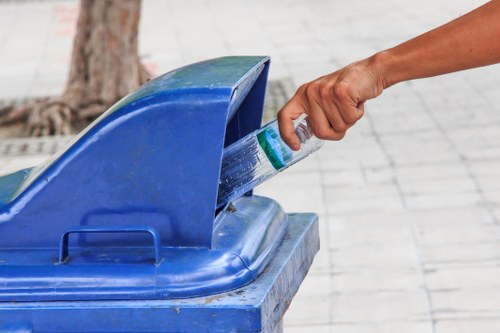 A person's hand is shown reaching into a blue outdoor rubbish bin, grasping a plastic bottle to place it inside for disposal. The bin is made of durable plastic with a textured surface and a curved, open-top lid. It is positioned on a paved area, possibly a sidewalk or courtyard, with a tree trunk visible in the background. The surrounding environment suggests an urban or town setting, with natural light illuminating the scene. This image is relevant to waste collection and rubbish removal services provided by companies such as Commercial Waste Holborn, which operate in the Holborn area. The focus is on environmentally conscious disposal practices, exemplifying proper waste segregation and recycling efforts typical of professional rubbish collection in London.